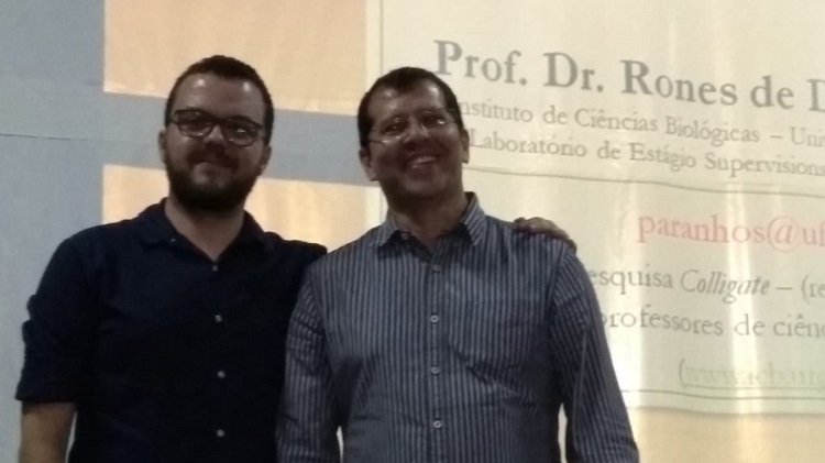 Paulo Henrique e Rones, de pé, sorrindo, Ambos de pele clara, cabelos escuros e óculos. Rones de camisa preta e Paulo, cinza