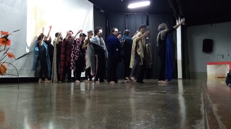 11 atores em pé, em fila indiana, todos com uma espécie de coberta ou lenço sobre seu corpo. Eles seguram, cada um, uma taça, braço que segura a taça erguido